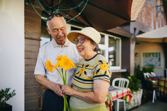 an older couple standing outside their house holding a bunch of yellow flowers
