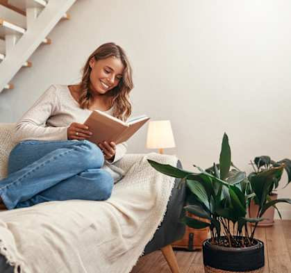 A woman is sitting on a couch reading a book with a lamp and a potted plant nearby.