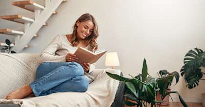 A woman is sitting on a couch reading a book with a lamp and a potted plant nearby.