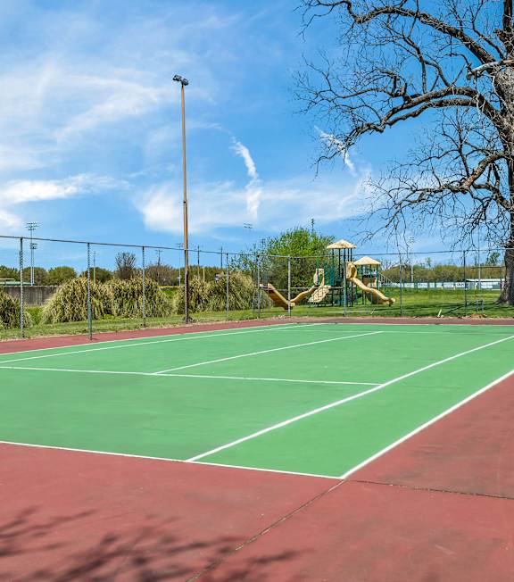 A tennis court with a green and red surface and a playground in the background.