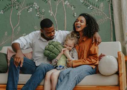 A family of three is sitting on a couch with a green wallpaper in the background.