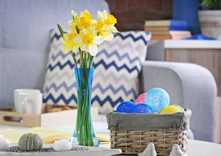 A vase with yellow flowers sits on a table next to a basket of colorful eggs.