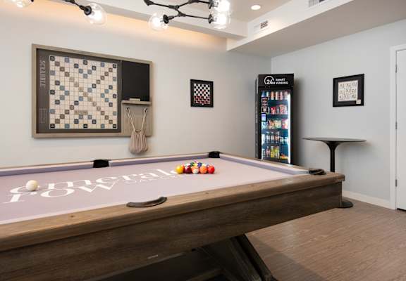 A pool table with a white cloth and balls in the foreground with a framed picture of a chess board on the wall behind it.