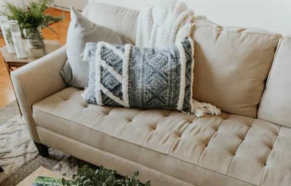 A beige couch with a grey and white throw pillow and a white blanket on it.