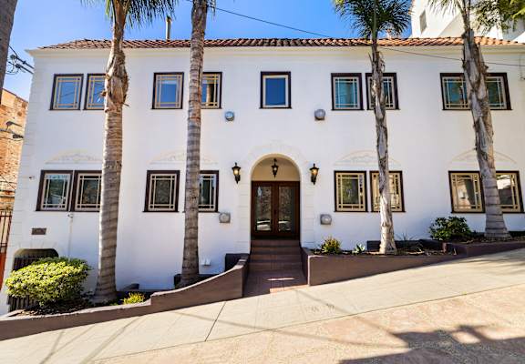 A white house with a brown door and windows surrounded by palm trees.