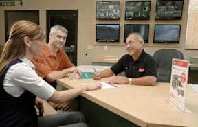 A man in a black shirt is sitting at a desk with two other people, in front of a wall with multiple monitors.