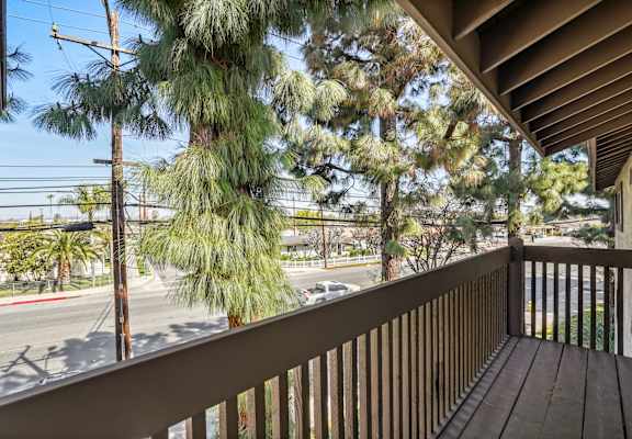 A balcony with a wooden floor and a railing overlooks a street with cars and trees.