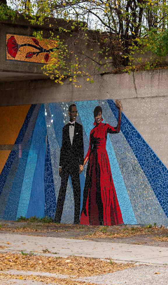 A mural of two people in formal attire is painted on a wall under an overpass.