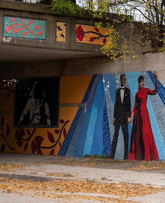 A mural of two people in formal attire is painted on a wall under an overpass.