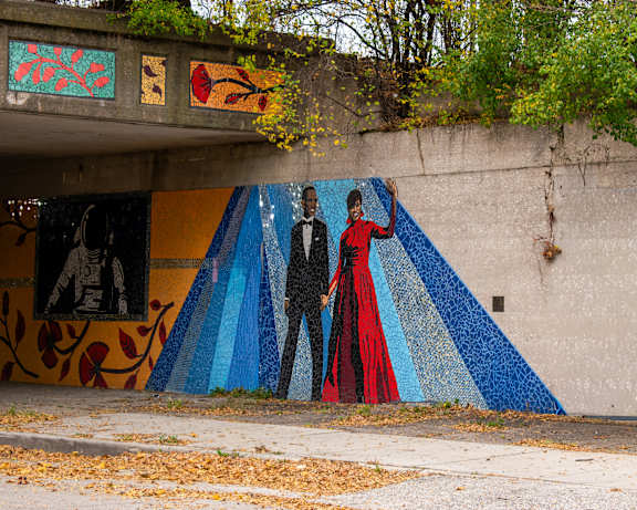 A mural of two people in formal attire is painted on a wall under an overpass.