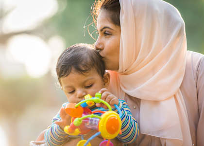 A woman in a light-colored hijab is kissing a baby while the baby is playing with a colorful toy.