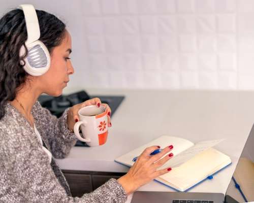 A woman with headphones on is reading a book while holding a mug.