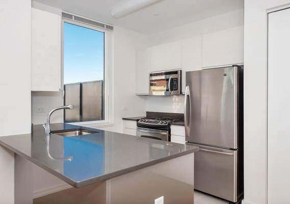 A modern kitchen with a stainless steel refrigerator and a sink with a blue tinted window at The Maximilian Apartments, Long Island City, NY, 11101