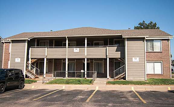 A black car is parked in a parking lot in front of a two-story apartment building.