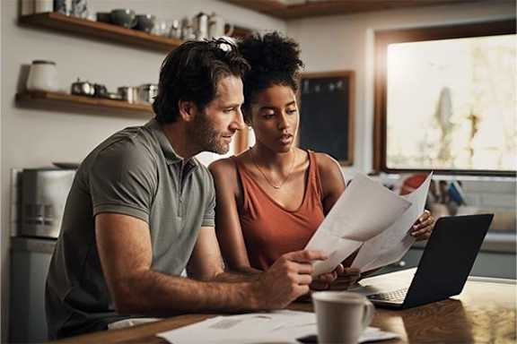 A man and woman are sitting at a table looking at papers and a laptop.