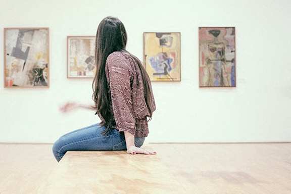 A woman in a pink shirt is sitting on the floor looking at paintings on the wall.