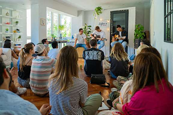 A group of people are sitting on the floor in a circle listening to a man playing guitar.