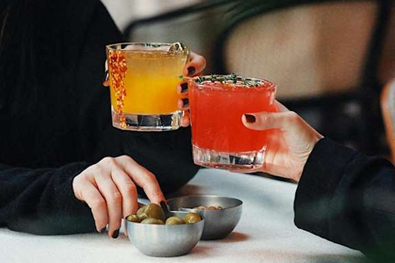 Two people toasting with drinks and olives on a table.