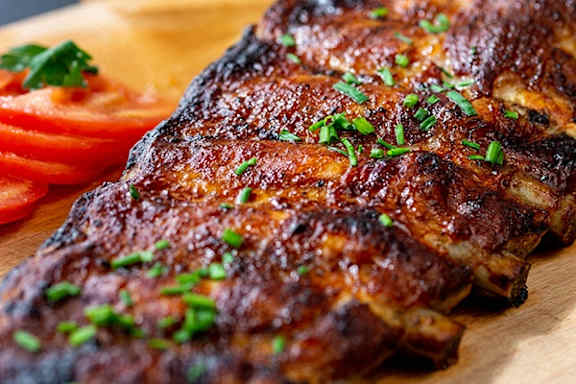 A wooden board holds a piece of grilled meat garnished with parsley and served with tomato slices.
