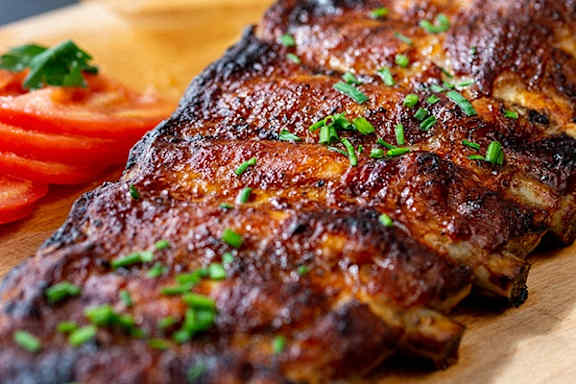 A wooden board holds a piece of grilled meat garnished with parsley and served with tomato slices.