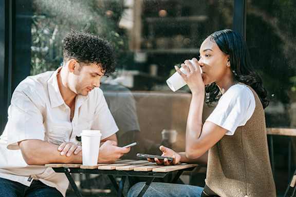 A man and a woman are sitting at a table, with the woman drinking from a white cup.