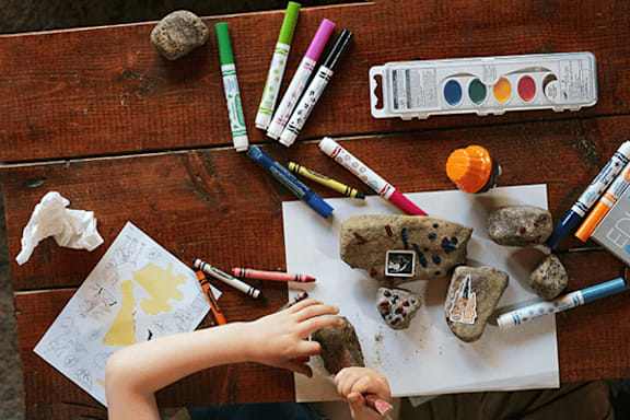 A child is painting rocks with various art supplies on a wooden table.