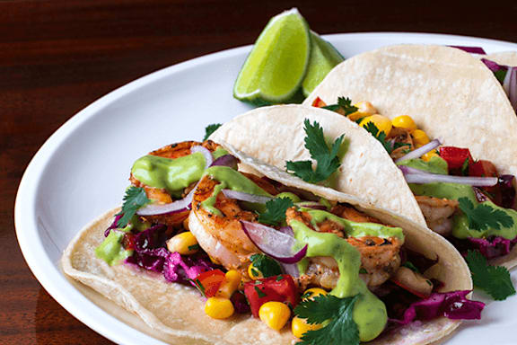 Three tacos with seafood, vegetables, and sauce on a white plate.