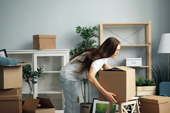A woman is moving boxes in a room with a white cabinet, a plant, and a wooden shelf.