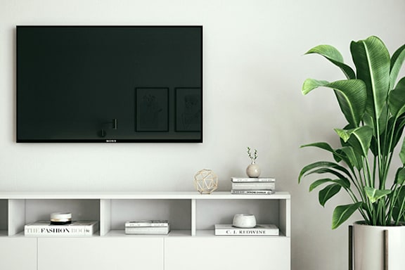 A black TV is mounted on a white wall above a white shelf with books and decorations.