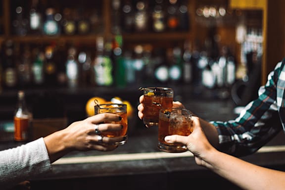 Two people toasting with drinks in front of a bar.