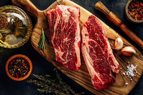 Two raw beef steaks on a cutting board with garlic, rosemary, and spices.
