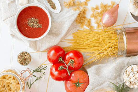 A bowl of tomato soup surrounded by ingredients like tomatoes, garlic, and spaghetti.