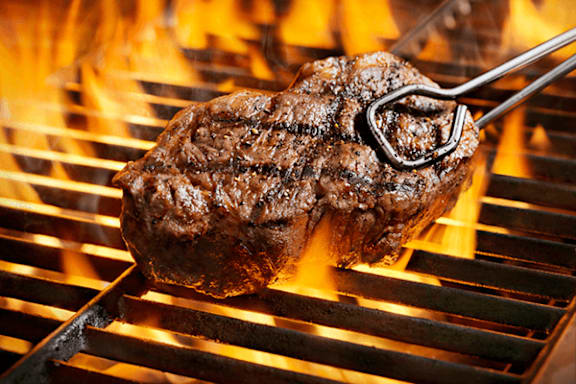 A steak is being cooked on a grill with visible flames.