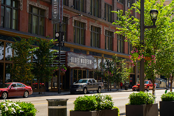A street view of a city with a red car and a blue car driving by a building with a sign that says "Blues Museum".