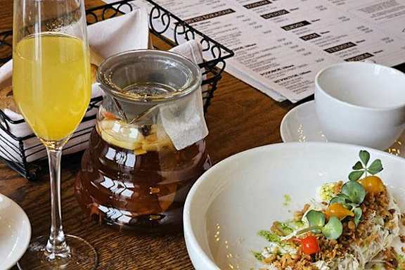 A glass of orange juice sits next to a bowl of food on a wooden table.