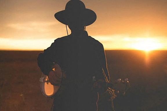 A silhouette of a cowboy holding a lasso stands in a field at sunset.