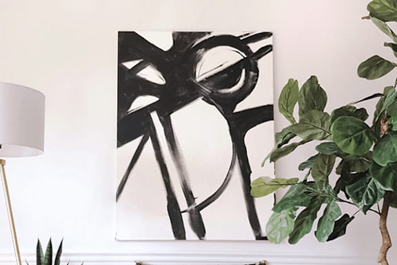 A black and white abstract painting hangs on a white wall above a green plant.