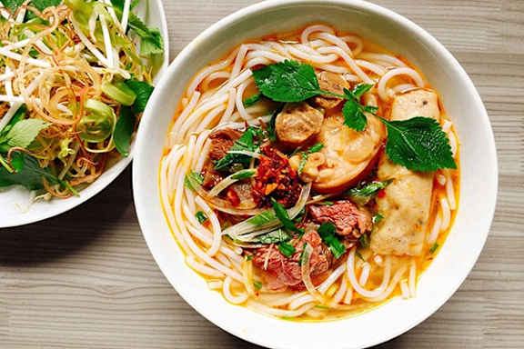 A bowl of noodles with meat and vegetables on a wooden table.