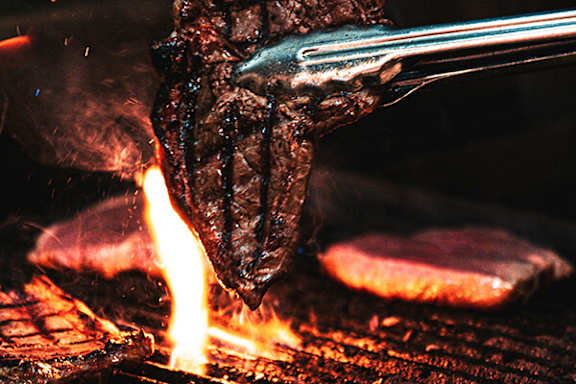 A steak is being cooked on a grill with flames visible.