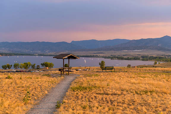 A gazebo sits in the middle of a field with a lake and mountains in the background.