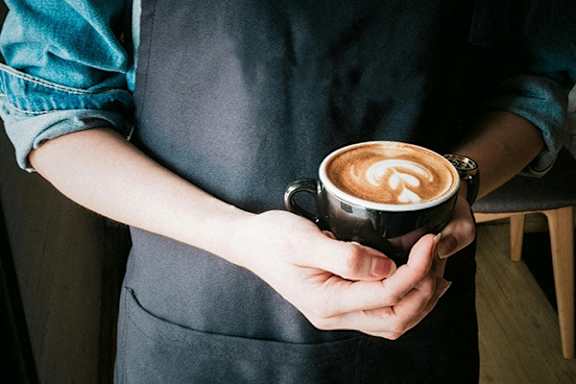 A person in a black apron holds a cup of coffee with a latte art design on top.