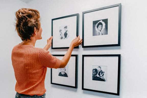 A woman in an orange top is hanging pictures on a wall.