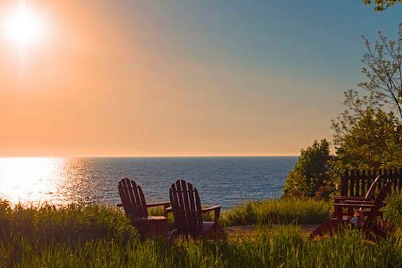 Two chairs are on a grassy hill overlooking the ocean at sunset.
