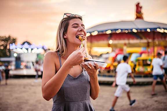 A woman eating a hot dog in front of a carnival ride.