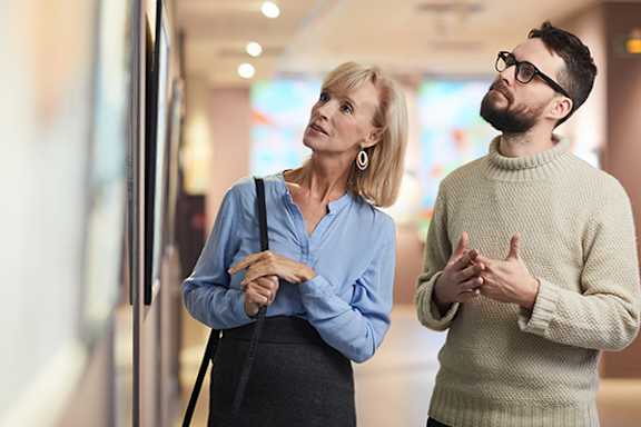 A man and a woman are standing in front of a painting, with the woman looking at it and the man gesturing with his hands.