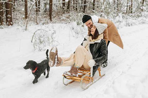 A man and woman sledding with a black dog.