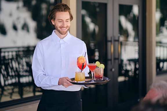 A man in a white shirt and black pants is holding a tray with drinks on it.