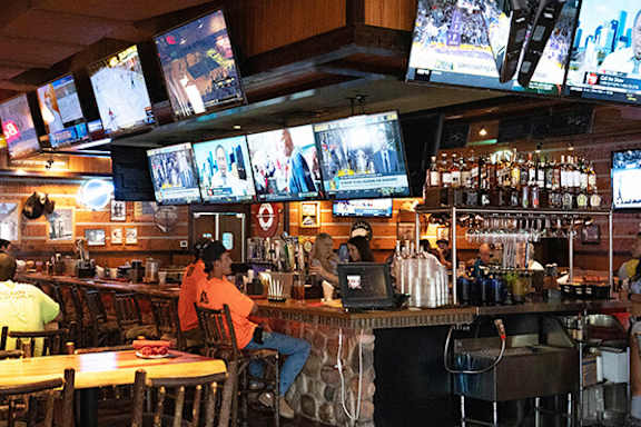 A man in an orange shirt is sitting at a bar with multiple televisions mounted on the wall.