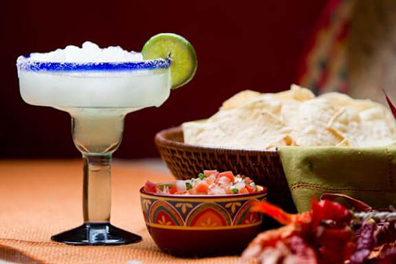 A margarita glass with a salted rim and a lime wedge on the rim, accompanied by a bowl of nachos and a basket of tortilla chips.