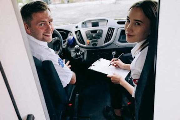 A man and a woman are sitting in a car, the man is driving and the woman is writing something on a piece of paper.
